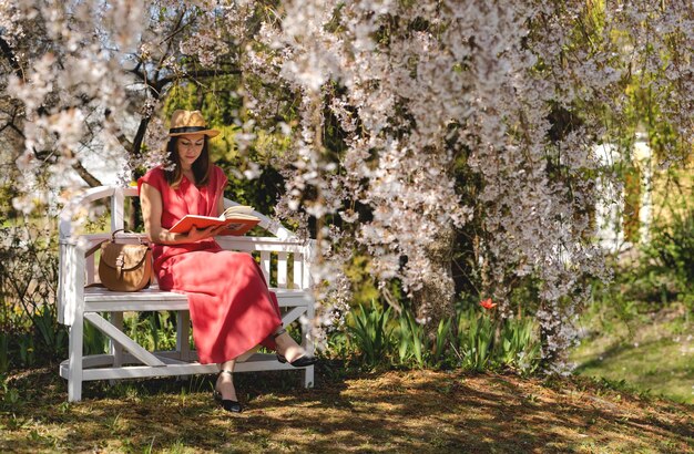 Una hermosa joven está sentada en un elegante banco en un jardín de primavera bajo una flor de cerezo y leyendo un libro