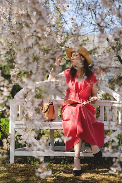 Una hermosa joven está sentada en un elegante banco en un jardín de primavera bajo una flor de cerezo y leyendo un libro