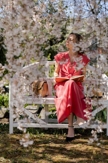 Una hermosa joven está sentada en un elegante banco en un jardín de primavera bajo una flor de cerezo y leyendo un libro