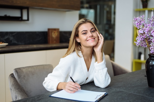 Una hermosa joven está sentada a la mesa de su cocina y escribe en un cuaderno. Hacer una lista de compras desde casa.