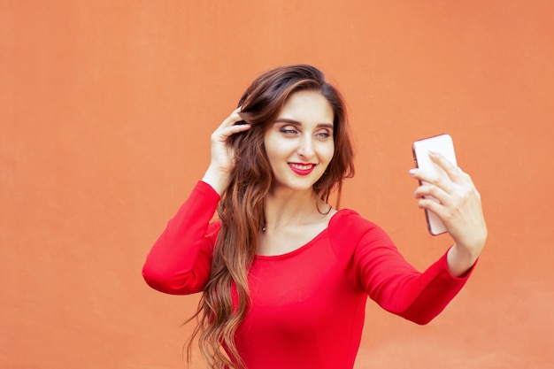 Hermosa joven está tomando selfie con smartphone sobre fondo naranja.