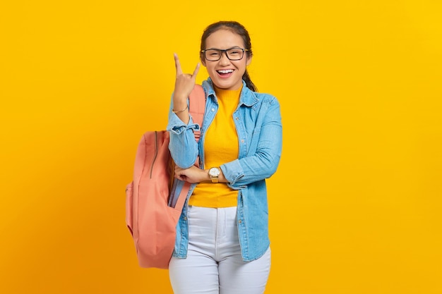 Hermosa joven estudiante asiática en traje de mezclilla con mochila gritando con expresión loca haciendo símbolo de rock con las manos arriba aislado sobre fondo amarillo Educación en concepto de colegio universitario