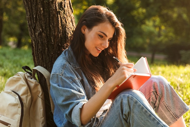 Hermosa joven estudiante escribiendo en un cuaderno mientras está sentado en el parque, apoyado en un árbol