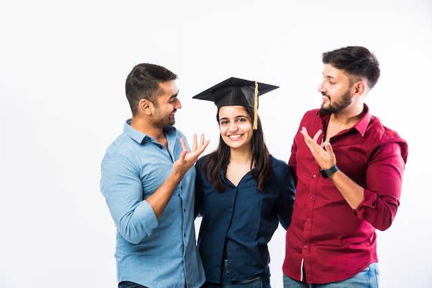 Hermosa joven estudiante india graduada celebrando el éxito con un amigo mientras lleva sombrero de ceremonia de graduación contra la pared blanca