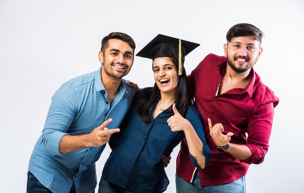 Hermosa joven estudiante india graduada celebrando el éxito con un amigo mientras lleva sombrero de ceremonia de graduación contra la pared blanca