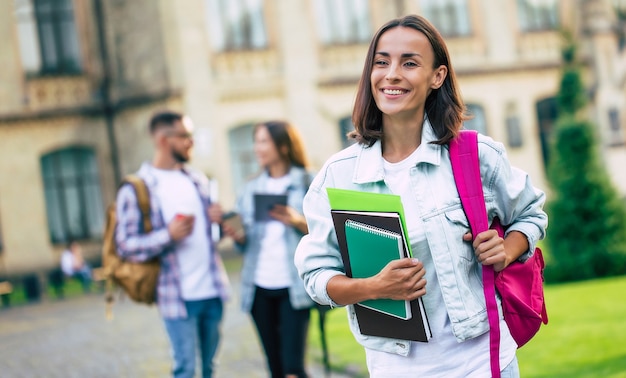 Hermosa joven estudiante morena en ropa de mezclilla con mochila y libros en las manos está de pie sobre un grupo de sus amigos estudiantes