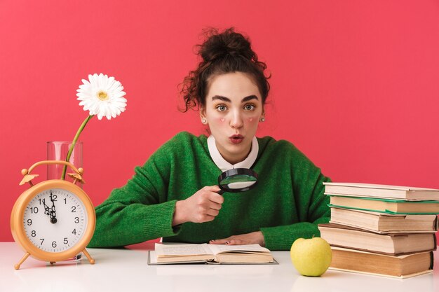 Hermosa joven estudiante nerd sentada en la mesa aislada, estudiando con libros, lupa