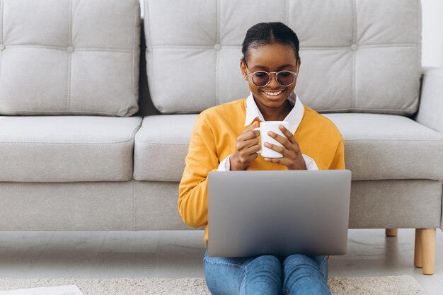 Hermosa joven estudiante universitaria negra sentada en el suelo, tomando café y haciendo un proyecto en una laptop