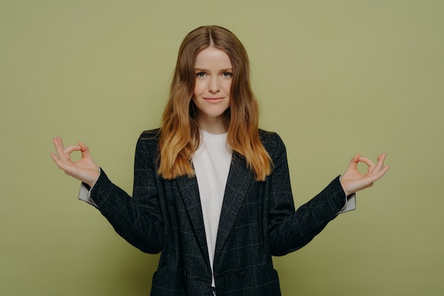 Hermosa joven estudiante vestida con elegante blazer a cuadros manteniendo las manos en gesto mudra y meditando, sintiéndose tranquila y feliz mientras practica la meditación, mirando a la cámara