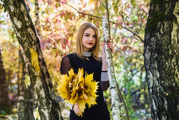 Hermosa joven estudiante vestida de negro con un ramo de hojas en el parque de otoño.