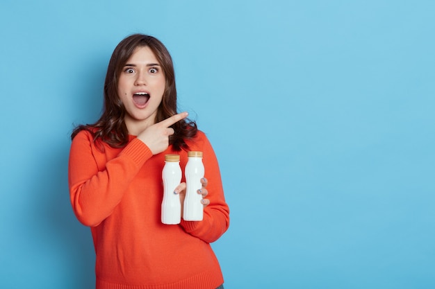 Hermosa joven europea sosteniendo dos botellas en blanco de leche o yogur en las manos mientras posa aislado en azul, sorprendida mujer señalando con el dedo