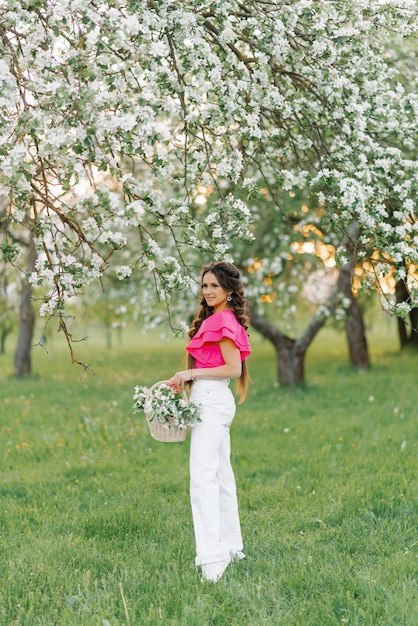 Una hermosa joven europea con trenzas en primavera en el parque sostiene una canasta de flores en un floreciente huerto de manzanos