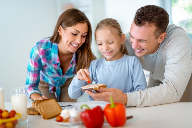 Hermosa joven familia preparando comida saludable para el desayuno en la cocina doméstica. La madre le enseña a la niña cómo hacer el sándwich y el padre mira lo que está haciendo.