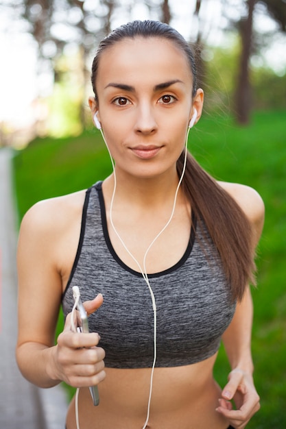 Hermosa joven feliz delgada para correr con un teléfono está escuchando música al aire libre en el parque