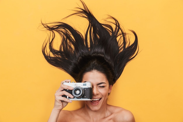 Hermosa joven feliz se encuentra aislado en la pared amarilla sosteniendo la cámara fotografiando