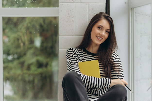 Una hermosa joven feliz está sentada en el alféizar de la ventana con un libro en las manos