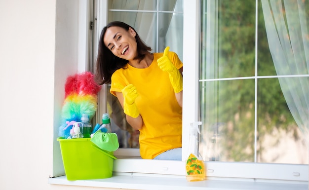 Hermosa joven feliz está usando un plumero y un spray mientras limpia una ventana en la casa