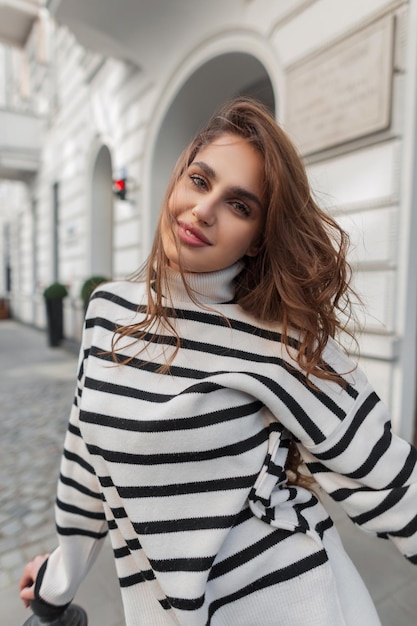 Hermosa joven feliz modelo morena con suéter a rayas de moda posando en la ciudad cerca de un edificio vintage blanco
