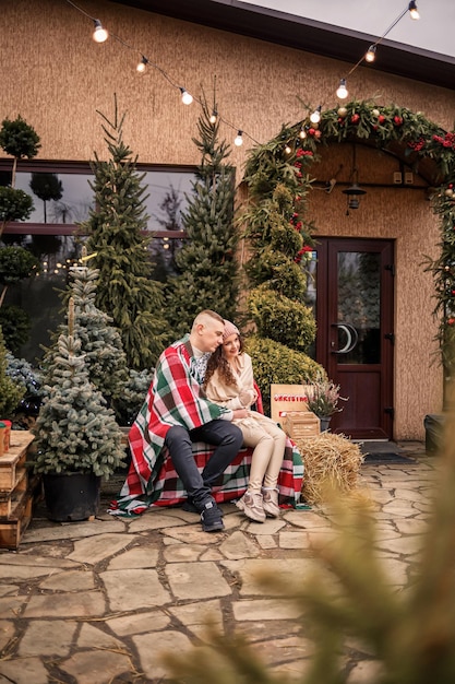 Hermosa joven feliz pareja sonriente envuelta en una manta escocesa Árboles de Navidad verdes en el fondo Las vacaciones de invierno Esperando el nuevo año