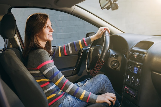 Foto una hermosa joven feliz sonriente europea de pelo castaño con piel limpia y saludable vestida con una camiseta a rayas se sienta en su coche con interior negro. concepto de viaje y conducción.