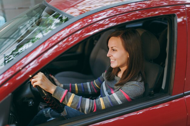 Una hermosa joven feliz sonriente europea de pelo castaño con piel limpia y sana vestida con una camiseta a rayas se sienta en su coche rojo con interior negro. Concepto de viaje y conducción.