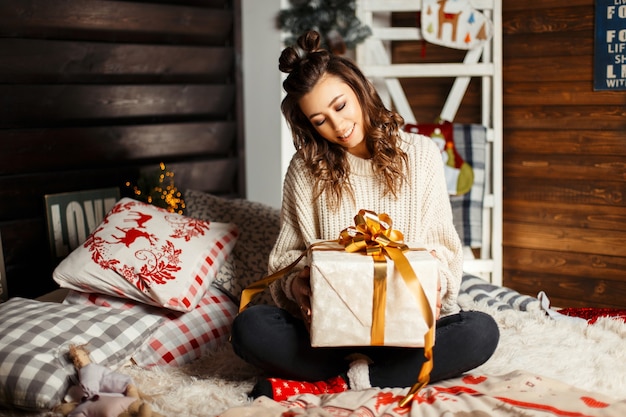 Hermosa joven feliz con una sonrisa en un suéter de punto vintage con un regalo en la cama en Nochebuena