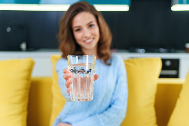 Foto hermosa joven feliz con un vaso de agua cristalina sentado en el sofá amarillo suave.