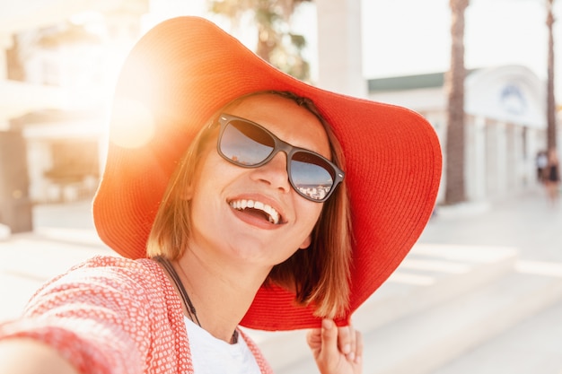 Foto hermosa joven felizmente sonríe al sol en un gran sombrero rojo brillante, concepto de vacaciones y viajes