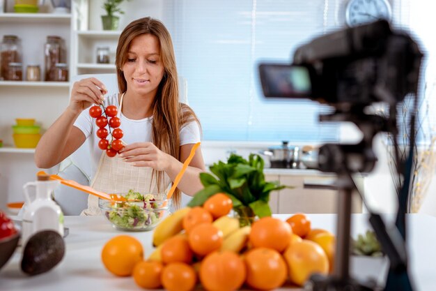 Hermosa joven filmando la transmisión de su blog sobre comida saludable en casa. Tiene tomate cherry en la mano y lo pone en una ensalada.