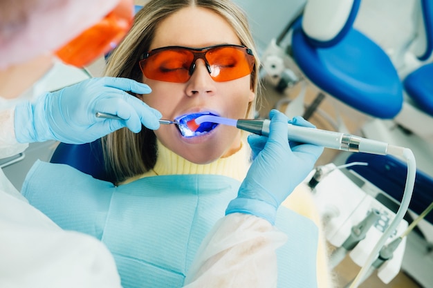 Una hermosa joven con gafas dentales trata sus dientes en el dentista con luz ultravioleta. relleno de dientes.