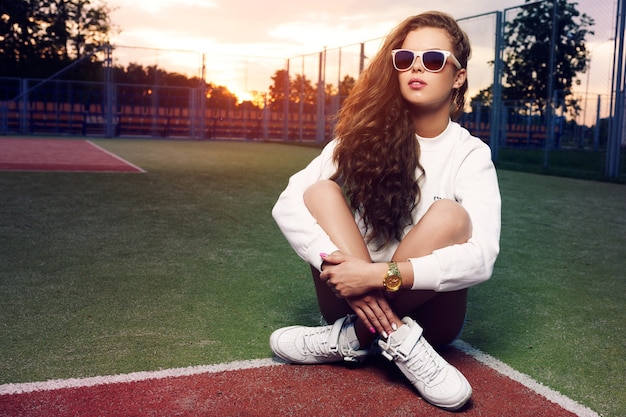 Hermosa joven con gafas de sol en la cancha de tenis. Hermoso cabello sano. Shorts de mezclilla. Zapatillas blancas