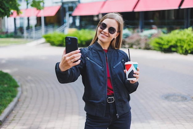 Hermosa y joven en gafas de sol escribe un mensaje en un teléfono celular y tomando café al aire libre