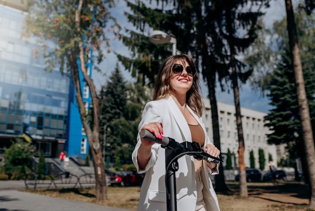 Hermosa joven con gafas de sol y traje blanco parada en su scooter eléctrico cerca de un edificio moderno y mirando hacia otro lado