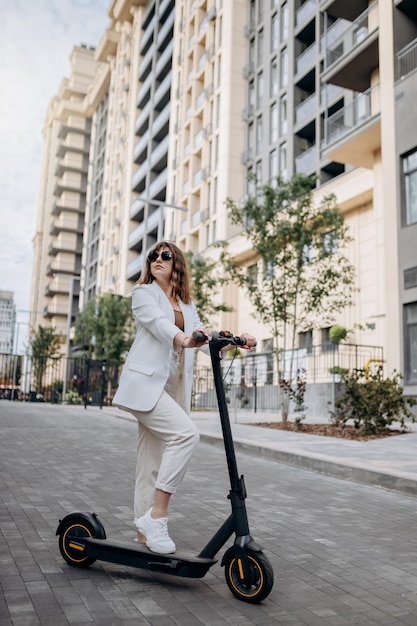 Hermosa joven con gafas de sol y traje blanco parada en su scooter eléctrico cerca de un edificio moderno y mirando hacia otro lado