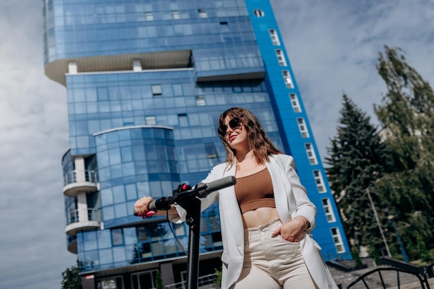 Hermosa joven con gafas de sol y traje blanco parada en su scooter eléctrico cerca de un edificio moderno y mirando hacia otro lado