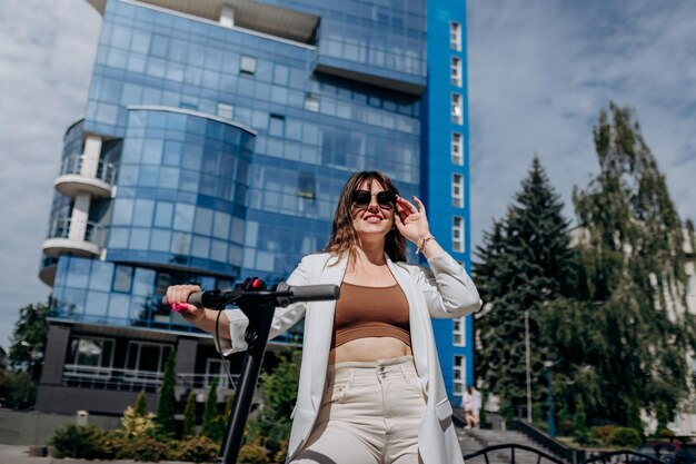 Hermosa joven con gafas de sol y traje blanco parada en su scooter eléctrico cerca de un edificio moderno y mirando hacia otro lado