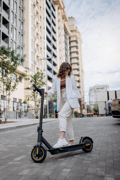 Hermosa joven con gafas de sol y traje blanco parada en su scooter eléctrico cerca de un edificio moderno y mirando hacia otro lado