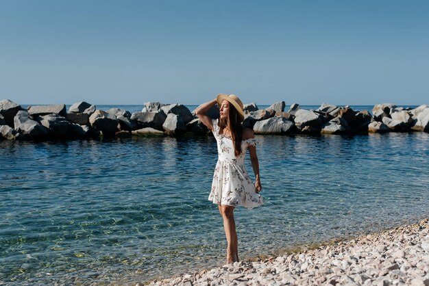 Una hermosa joven con gafas de sombrero y un vestido ligero camina a lo largo de la orilla del océano contra el fondo de enormes rocas en un día soleado Turismo y viajes turísticos