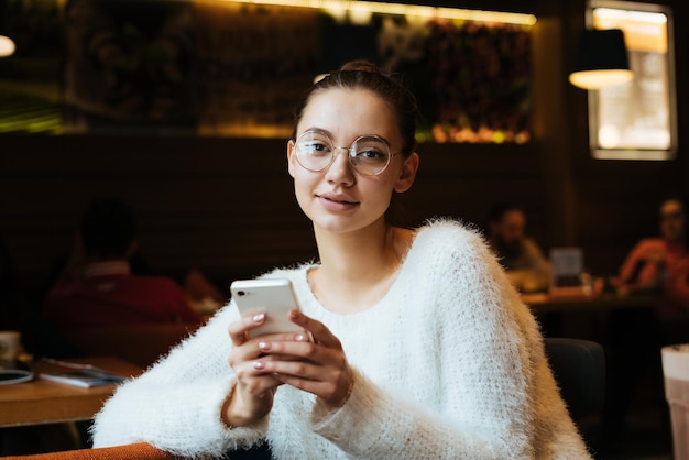 Hermosa joven con gafas y un suéter blanco sentada en un acogedor café sostiene un teléfono inteligente y sonríe