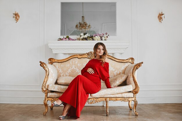 Hermosa joven gestación en vestido de noche rojo de moda se sienta en el sofá vintage dorado en un interior de lujo decorado con flores