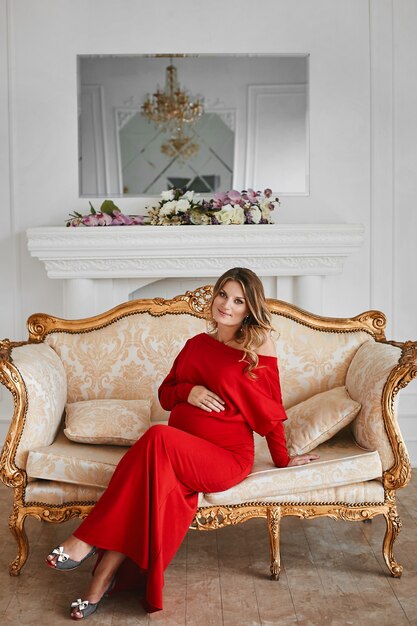 Hermosa joven gestación en vestido de noche rojo de moda se sienta en el sofá vintage dorado en un interior de lujo decorado con flores