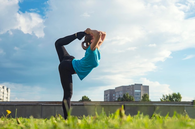 Hermosa joven gimnasta de pie con su pie sobre su cabeza en una de las poses de yoga en el parque de la ciudad en el césped