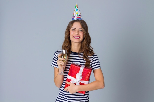 Hermosa joven con gorra de celebración sosteniendo un regalo rojo y una copa de champán