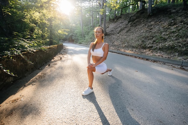 Una hermosa joven hace estocadas y calentamiento antes de correr el entrenamiento, en la carretera en un denso bosque, durante la puesta de sol. Un estilo de vida saludable y correr al aire libre.