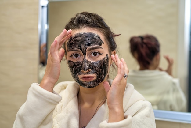 Hermosa joven haciendo tratamientos de belleza con máscara negra en la cara del acné Concepto de cuidado de la piel facial