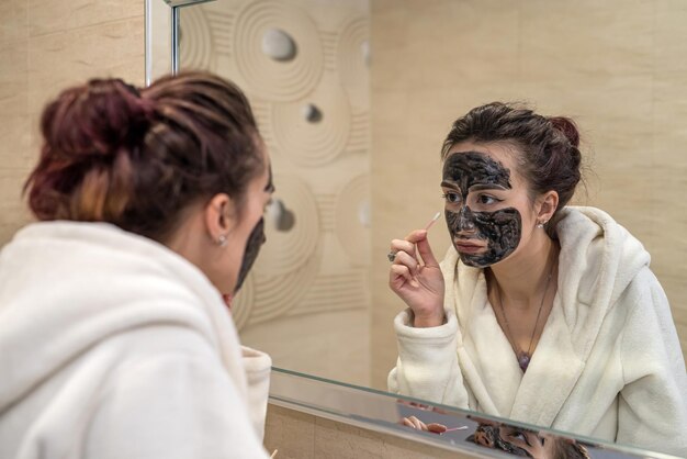 Hermosa joven haciendo tratamientos de belleza con máscara negra en la cara del acné Concepto de cuidado de la piel facial