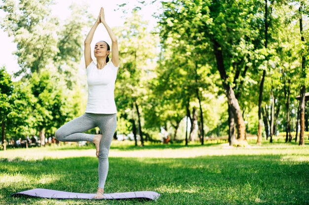 Hermosa joven haciendo yoga y de pie en la postura del árbol en la estera mientras se relaja y medita rodeado de naturaleza en el parque