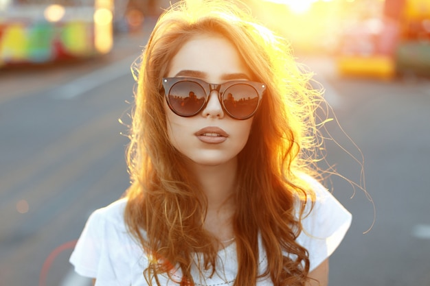 Hermosa joven hipster en gafas de sol de moda de pie en la playa en la increíble puesta de sol