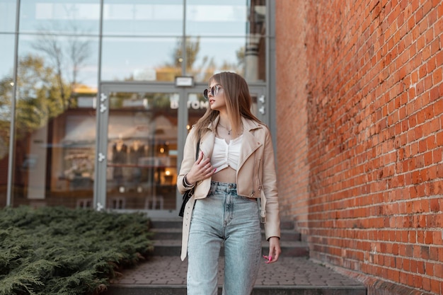 Hermosa joven hipster de moda con una chaqueta de cuero de moda con jeans de cintura alta y elegantes gafas de sol frescas caminando por la calle cerca del centro comercial