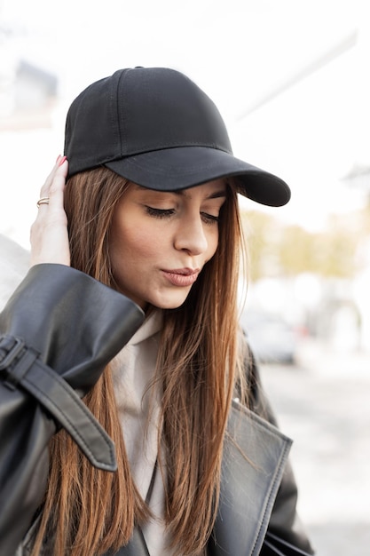 Hermosa joven hipster de moda con una gorra negra de moda y un abrigo de  cuero negro con una sudadera con capucha camina por la ciudad elegante  retrato de una mujer